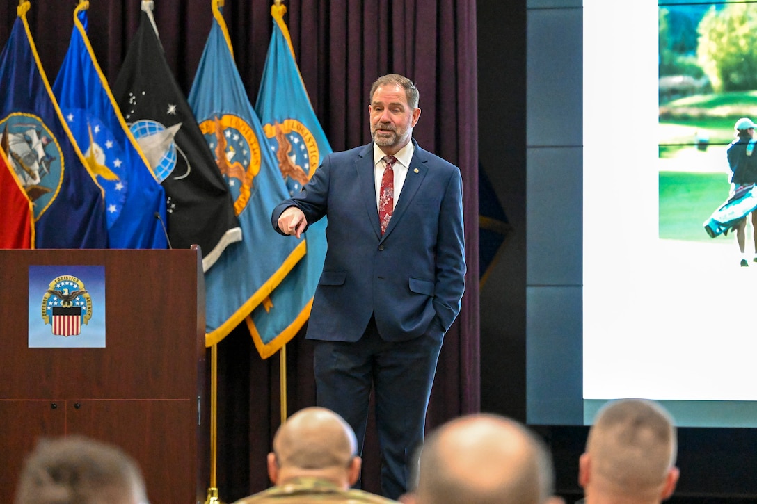 A man dressed in a dark suite stands on a stage talking to people in military uniforms.