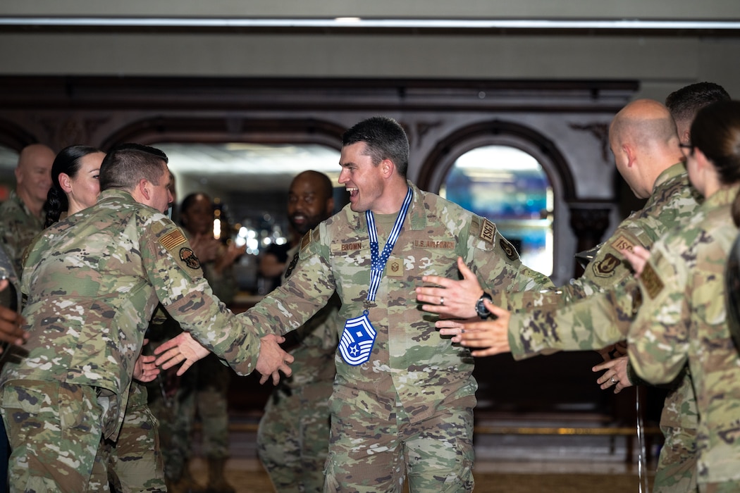 A smiling man, wearing military camouflage uniform, give high fives to other military members. A large blue and white rank insignia hangs around his neck