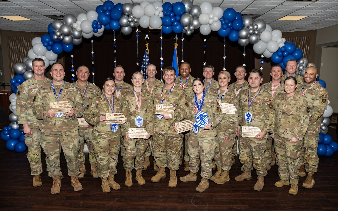 A group photo of men and women, wearing military camouflage uniforms, pose for a group photo with large SMSgt ranks hung from their necks and wooden plaques in hand