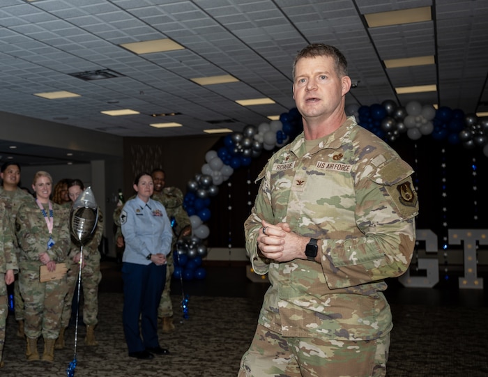 A man, wearing Air Force camouflage uniform, speaks in front of a crowd