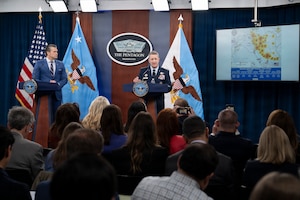 The secretary of war and the chairman of the Joint Chiefs of Staff stand at podiums on a stage while speaking to a seated audience.