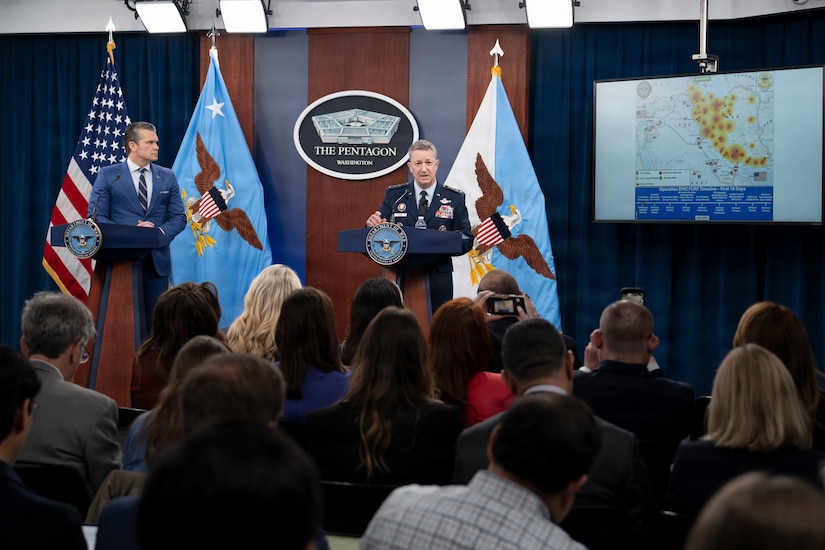 The secretary of war and the chairman of the Joint Chiefs of Staff stand at podiums on a stage while speaking to a seated audience.