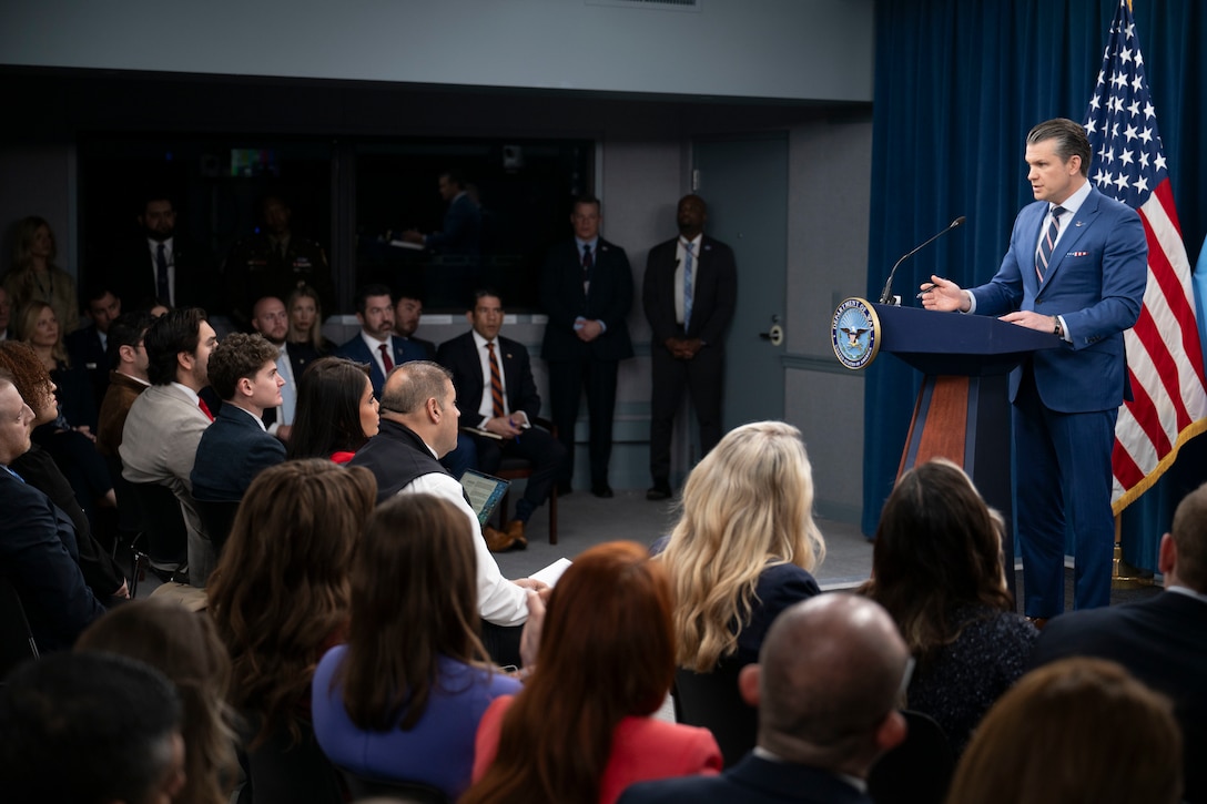 A man in business attire stands behind a lectern and speaks to a seated audience; behind him is an American flag.