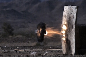 A small circle of black smoke radiates from a military round that explodes near a target on a range.