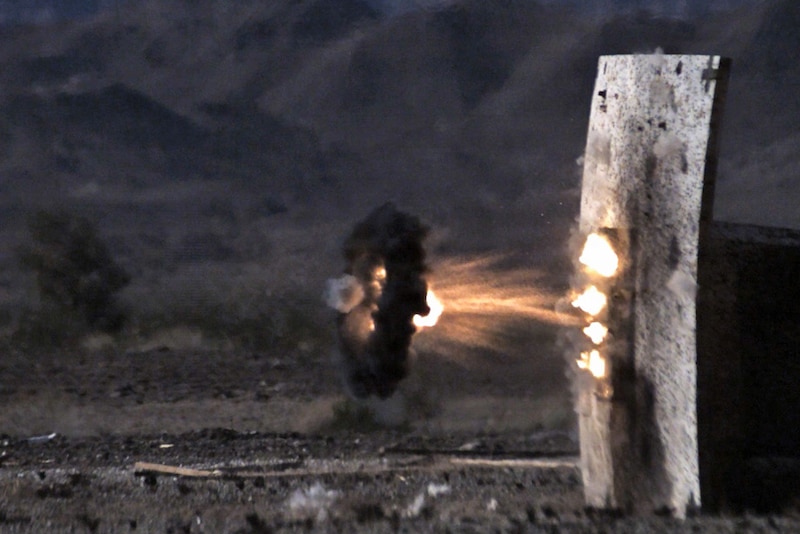 A small circle of black smoke radiates from a military round that explodes near a target on a range. 