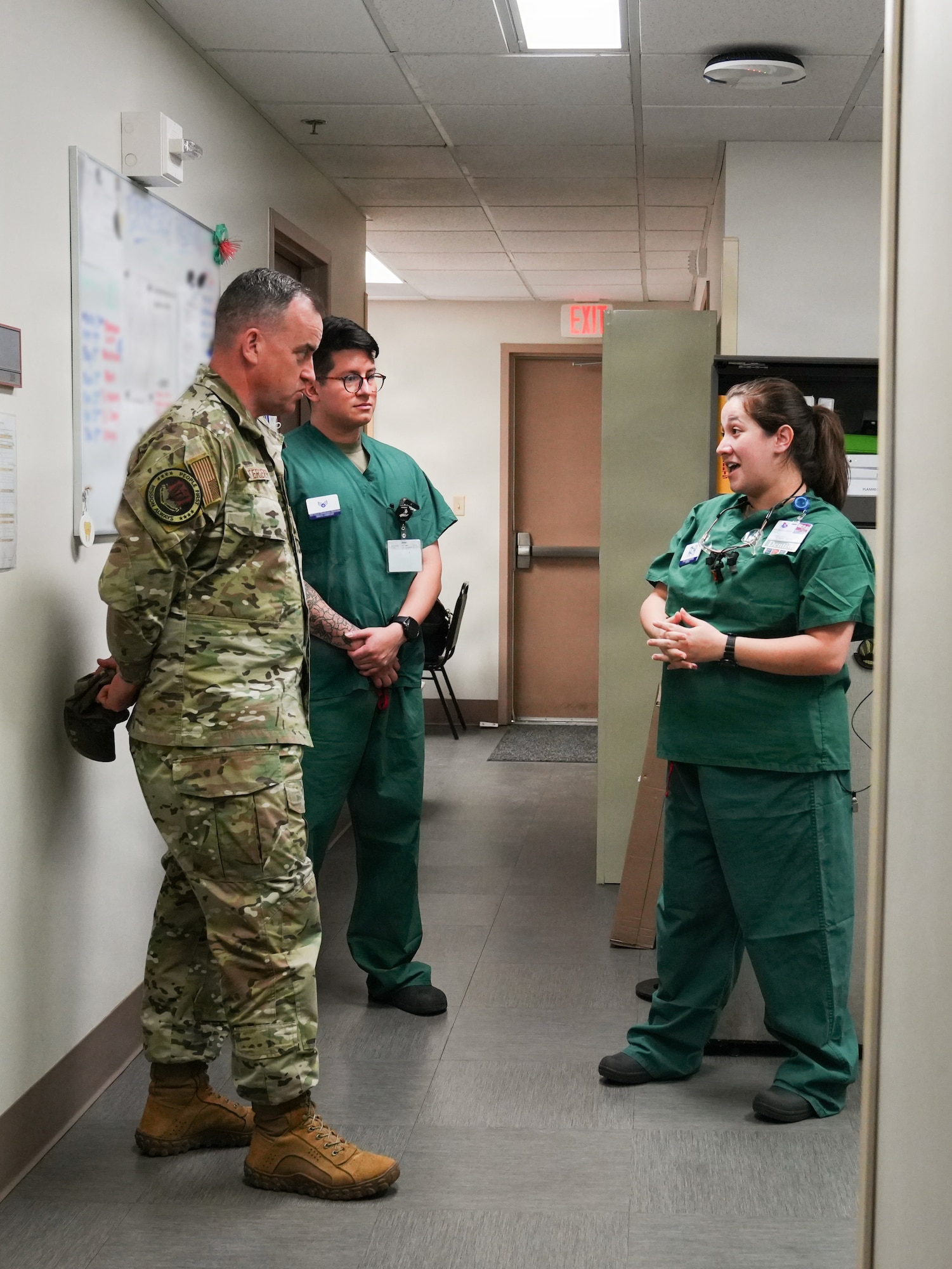A man in a military uniform stands in a hallway speaking to two people in medical scrubs.