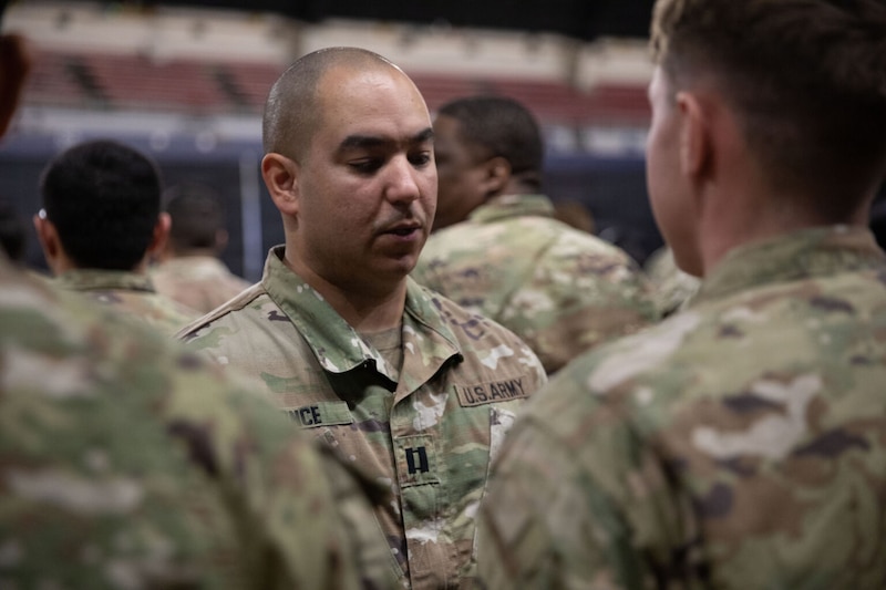 A man wearing a military camouflage uniform stands and looks down. Other men in the foreground stand at attention. 