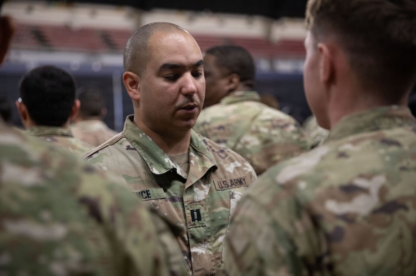 A man wearing a military camouflage uniform, stands while looking down. Other men in the foreground stand at attention.