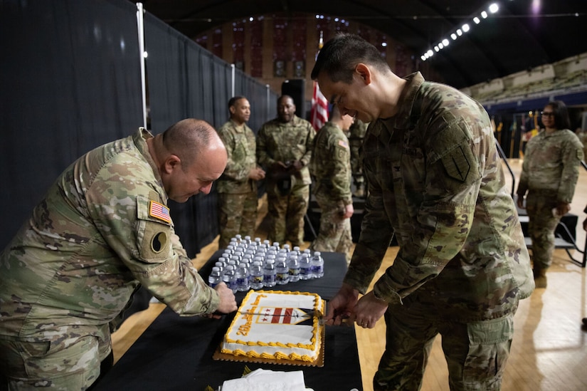 A man dressed in a camouflage military uniform cuts a cake as another man in similar attire looks on.