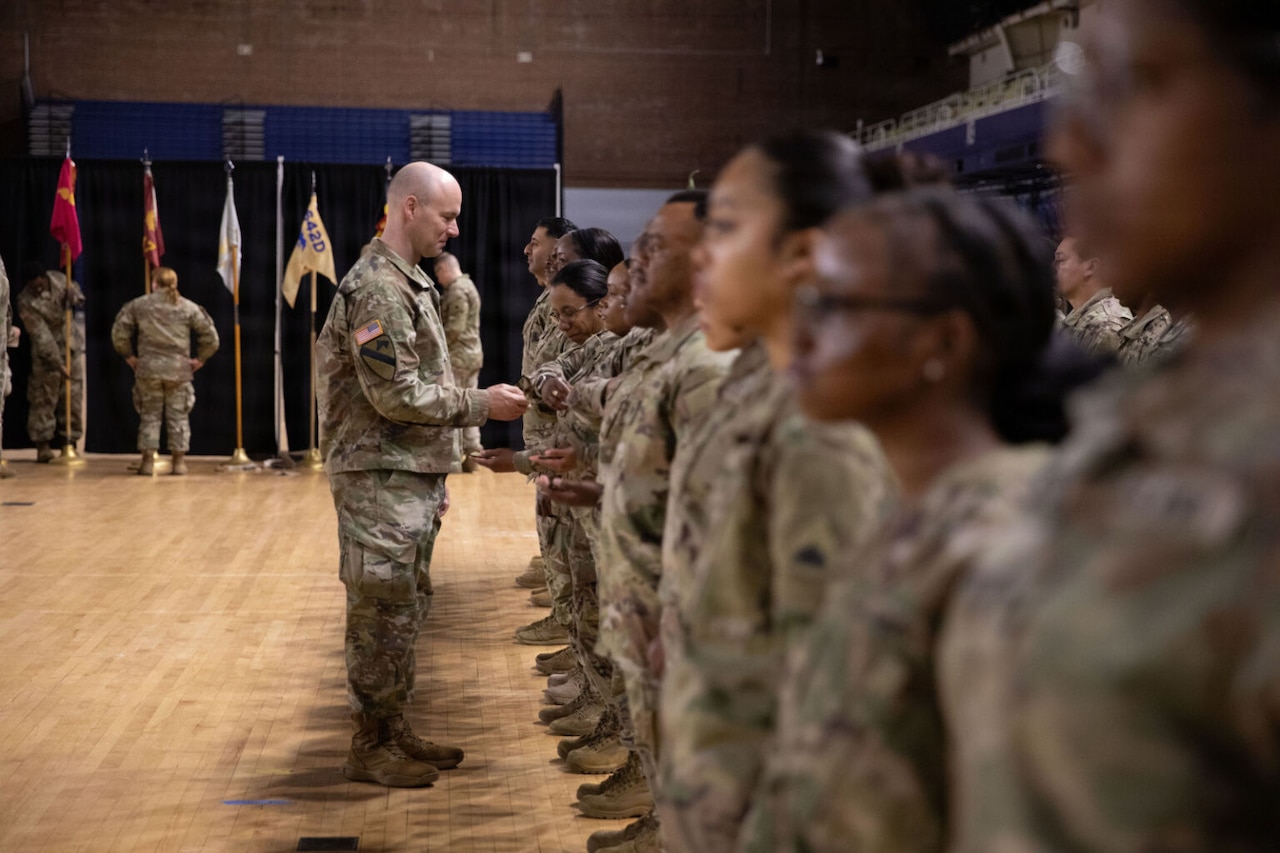 A group of people dressed in camouflage military uniforms stand at attention as a man also dressed in similar attire hands them a coin.