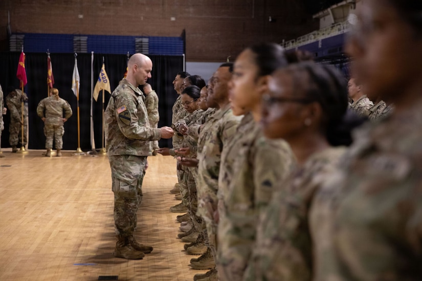 A group of people dressed in camouflage military uniforms stand at attention as a man also dressed in similar attire hands them a coin.