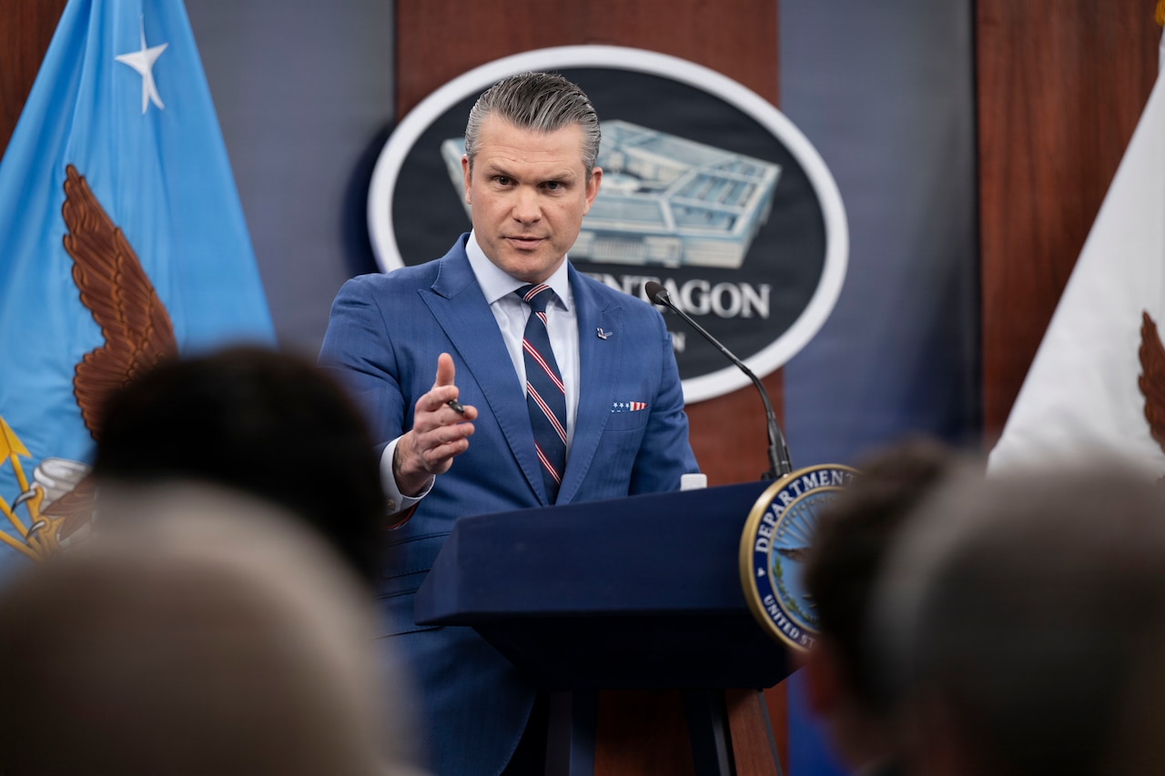 A man in business attire stands behind a lectern and speaks to a seated audience; behind him are a blue flag with an eagle in the center and a sign featuring a five-sided building with the word "Pentagon" written underneath it.