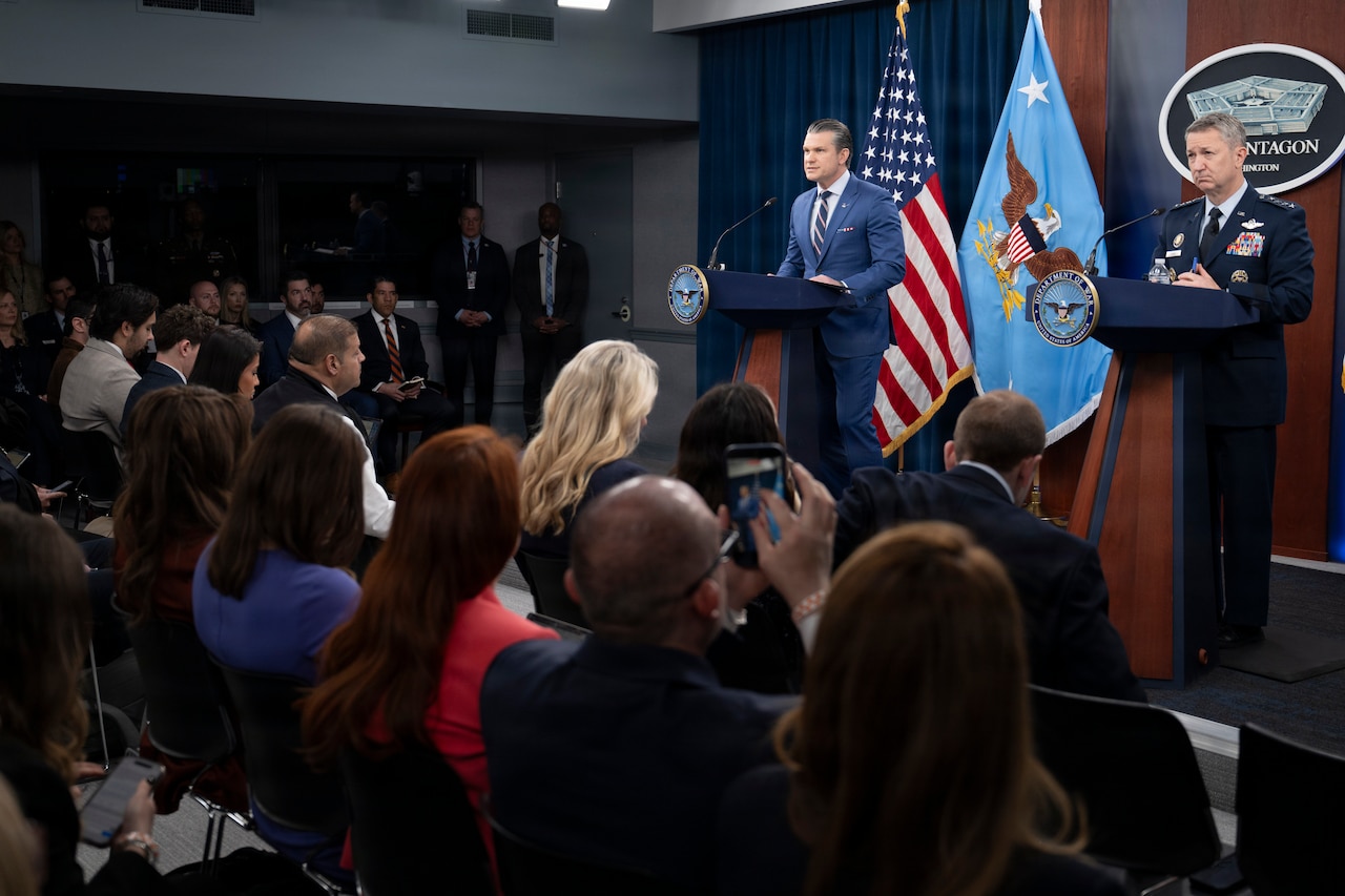 Two men, one in a suit and the other in a military uniform, stand behind lecterns on a stage. Behind them are two flags and a sign reading "The Pentagon." In front, a dozen people are seated.