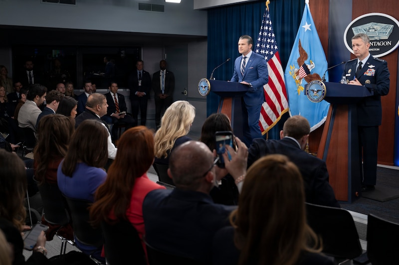 Two men, one in a suit and the other in a military uniform, stand behind lecterns on a stage. Behind them are two flags and a sign reading 'The Pentagon.' In front, a dozen people are seated.