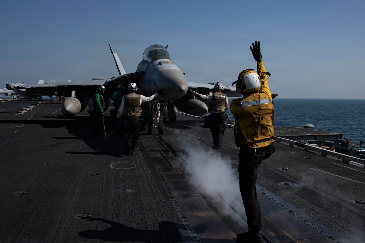 An aircraft sits on a flight deck of a large military ship. People in aviation gear prepare to launch it.