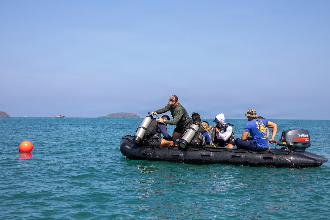 Navy divers from multiple nations operate a small rubber boat near a large orange ball in blue water.