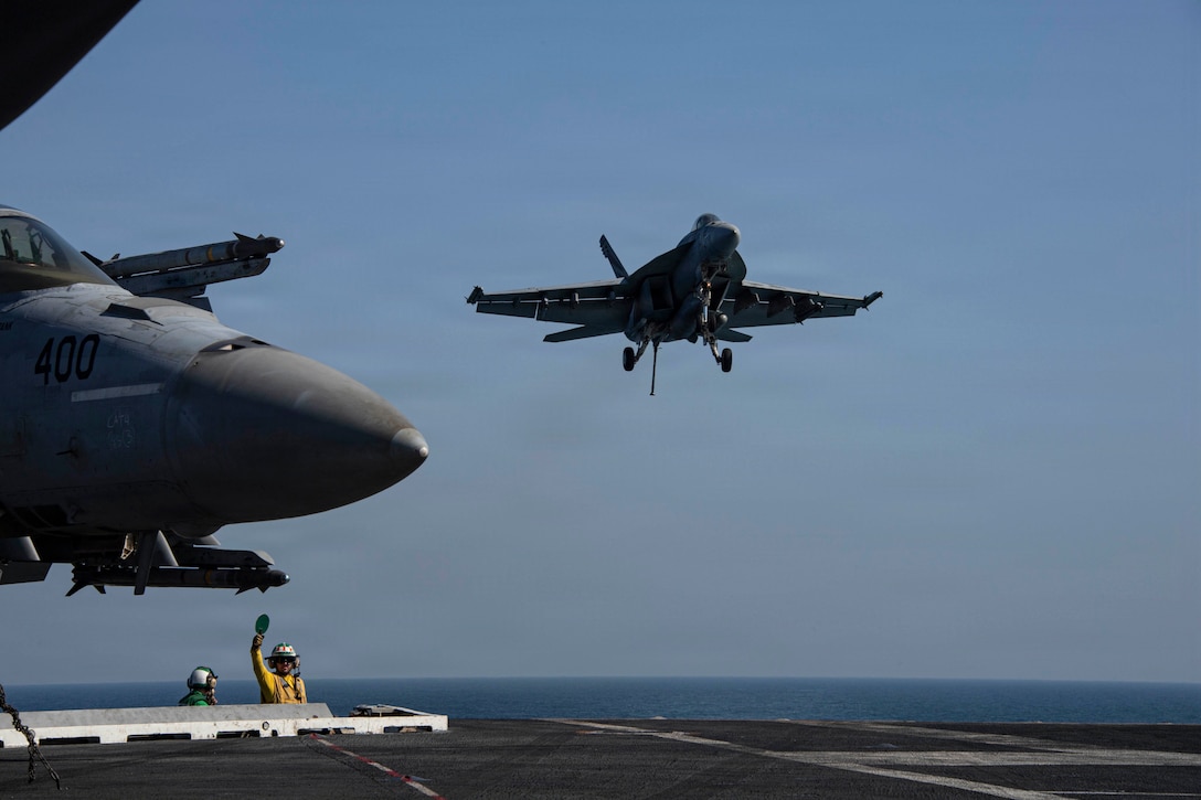 An aircraft in a blue sky prepares to land on a ship at sea as two sailors stand in the background near a parked aircraft on a tarmac.