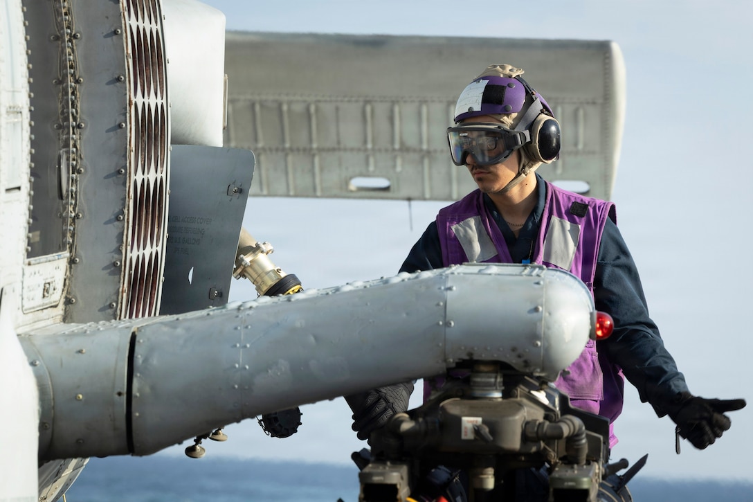A sailor in a purple vest connects a fuel hose to a partially visible helicopter aboard a ship at sea during the day.
