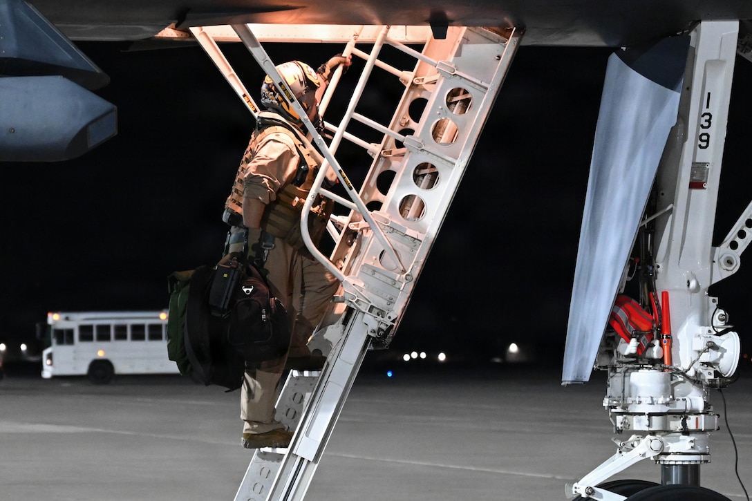 An airman climbs the ladder of an aircraft while carrying a large black bag in the dark.