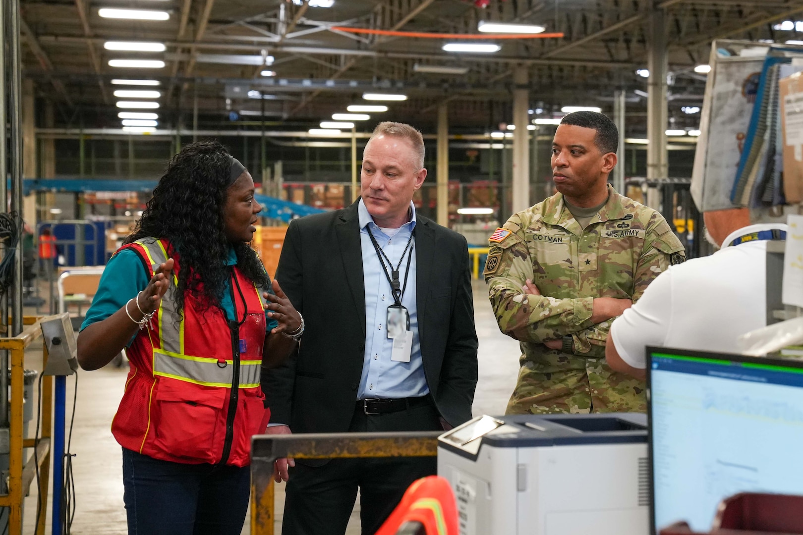 A man wearing a camouflage military uniform stands inside a large warehouse speaking to a man and woman.