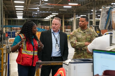 A man wearing a camouflage military uniform stands inside a large warehouse speaking to a man and woman.