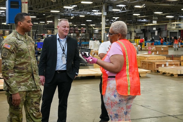 A man wearing a camouflage military uniform stands inside a large warehouse speaking to a man and woman.