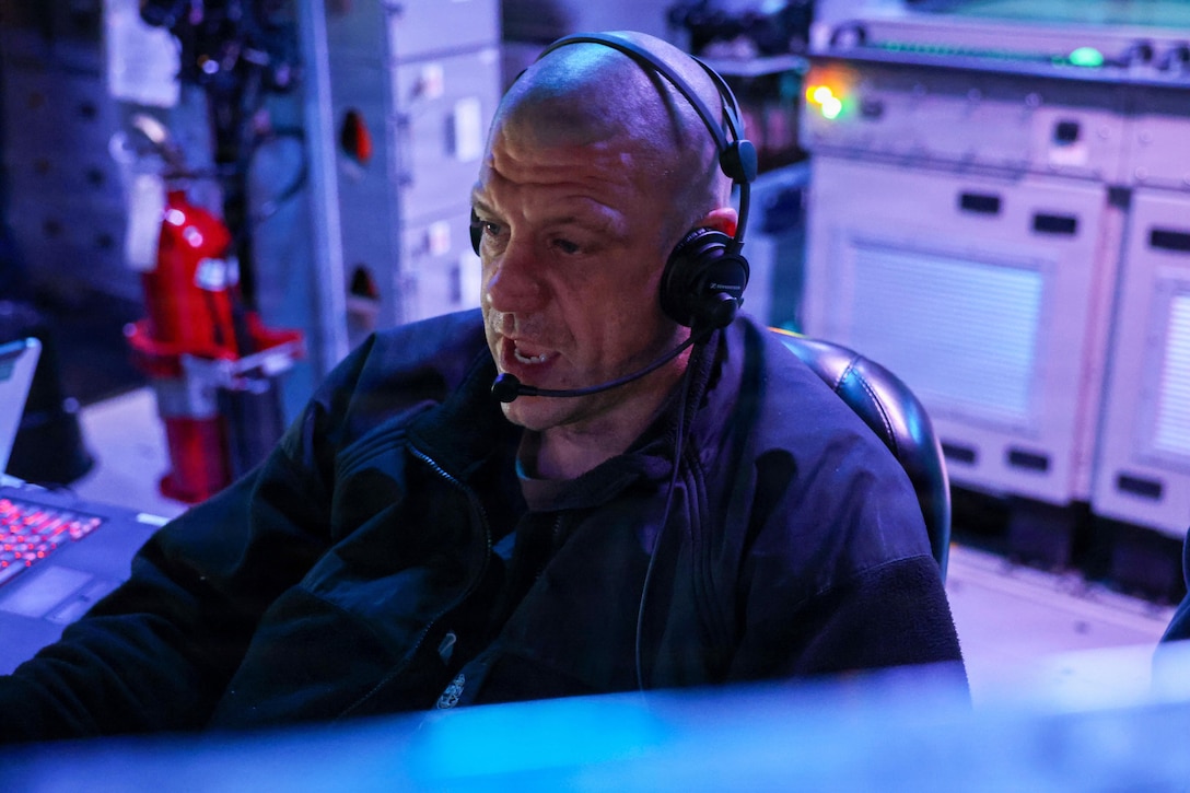 A sailor wearing a headset in a dark information center illuminated by blue light.