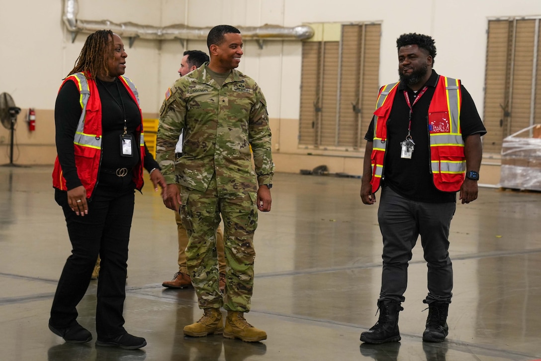 A man wearing a camouflage military uniform stands inside a large warehouse speaking to a man and woman who are wearing orange safety vests.