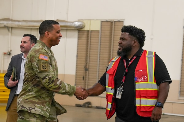 A man wearing a camouflage military uniform shakes another man's hand who is wearing an orange safety vest as they stand inside a large warehouse.