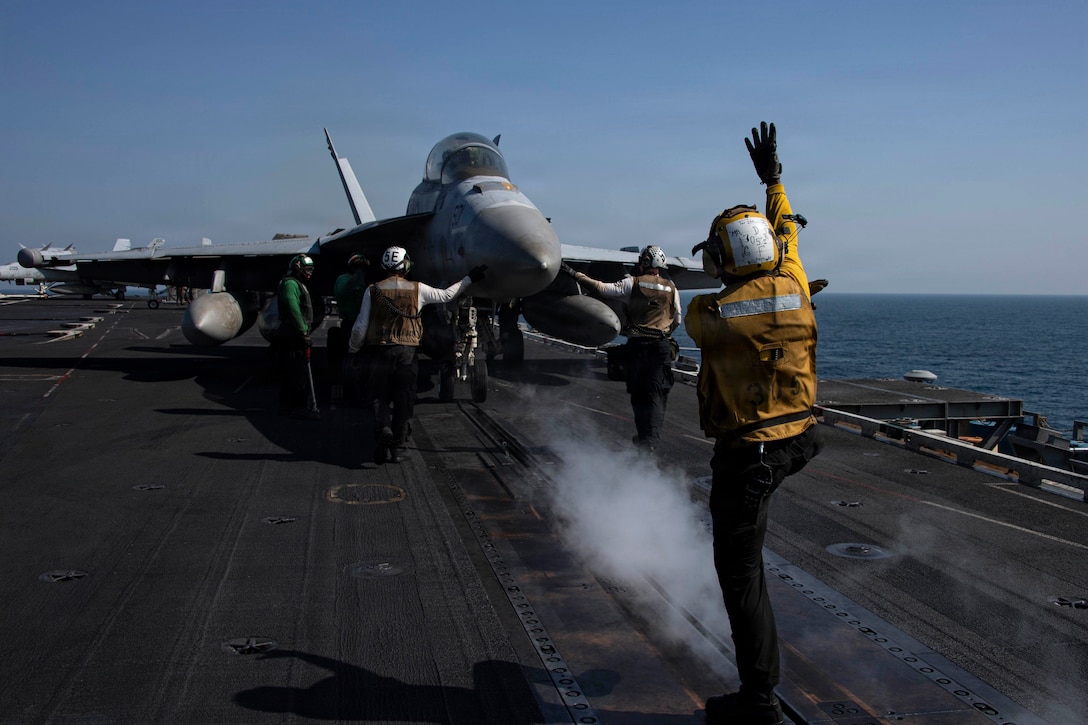 A sailor signals in front of an aircraft as three fellow sailors gather around it aboard a ship at sea under a blue sky.