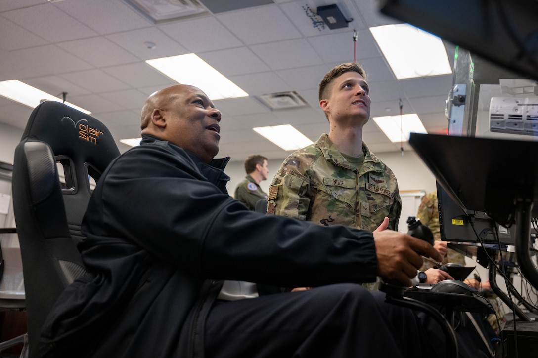 Two men look at a screen, as one holds a joystick.