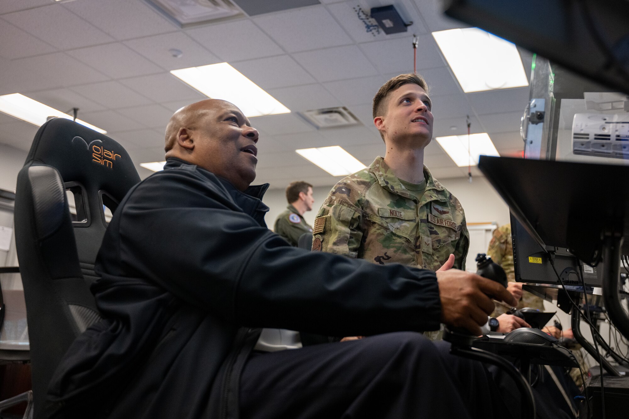 Two men look at a screen, as one holds a joystick.
