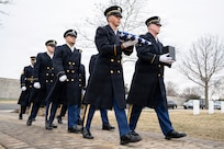 A group of Army Soldiers in ceremonial uniforms are marching in two rows during a funeral at a cemetery, with one at the front carrying an urn and another carrying a folder U.S. flag