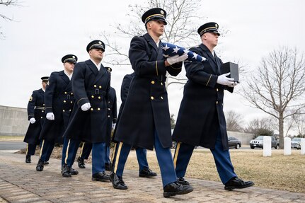 A group of Army Soldiers in ceremonial uniforms are marching in two rows during a funeral at a cemetery, with one at the front carrying an urn and another carrying a folder U.S. flag