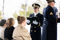 An Army Soldier in ceremonial uniform with white scarf and white gloves is holding a book while speaking to people who are seated during a funeral.