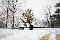An Army soldier in ceremonial uniform with red hat is seen in the distance performing on a bugle in the middle of a cemetery.