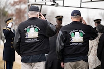 Two men wearing black jackets that have the words Guard of Honor about a graphic image are saluting away from the camera during a funeral.