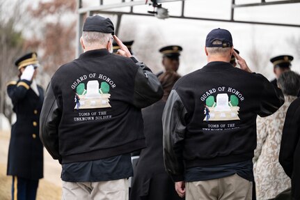 Two men wearing black jackets that have the words Guard of Honor about a graphic image are saluting away from the camera during a funeral.