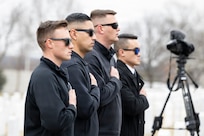 Four men wearing black sunglasses and black fleece sweaters are standing in a row with their hands on their hearts facing to the right, and there is a camera on a stand in the background.