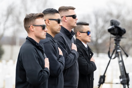 Four men wearing black sunglasses and black fleece sweaters are standing in a row with their hands on their hearts facing to the right, and there is a camera on a stand in the background.