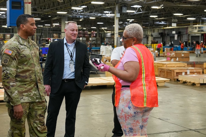 A man wearing a camouflage military uniform stands inside a large warehouse speaking to a man and woman.