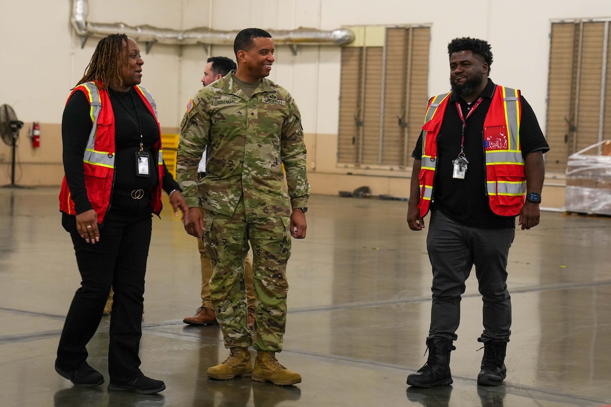 A man wearing a camouflage military uniform stands inside a large warehouse speaking to a man and woman who are wearing orange safety vests.
