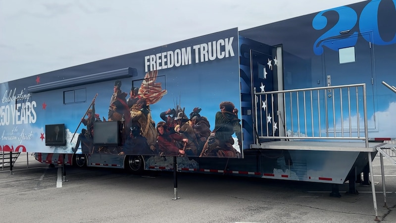 A trailer with 'Freedom Truck' written on the side sits in a parking lot. A historic battle scene is painted on the side.