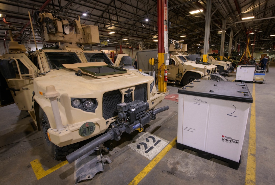 Several tan-colored armored vehicles are shown in a large factory during the manufacturing process.