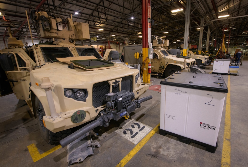 Several tan-colored armored vehicles are shown in a large factory during the manufacturing process.