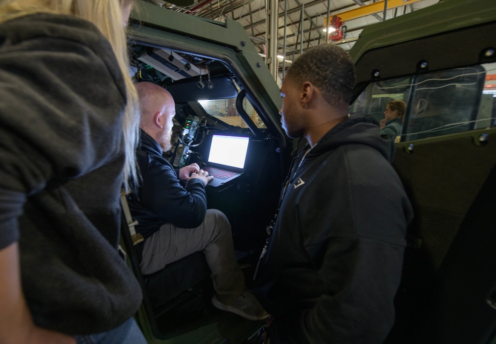Two people look on as a man sits inside a tactical vehicle, working on an illuminated screen in the dashboard.