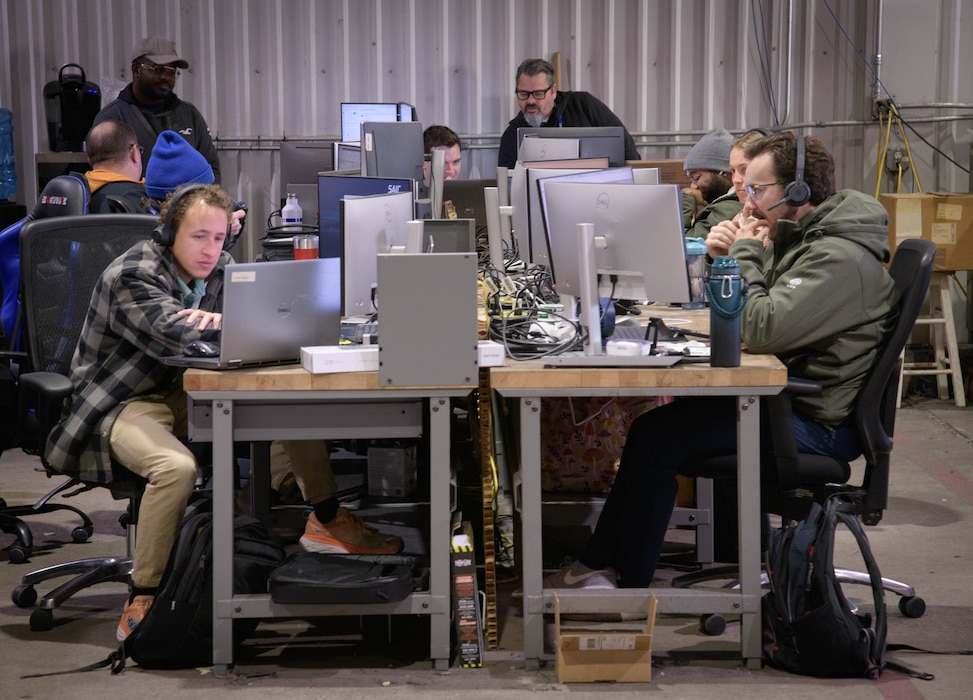 Several men sit at shared wooden desks in an industrial-style office, working intently on their laptops and monitors.