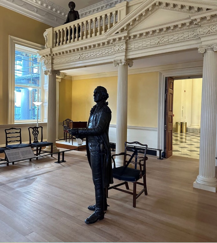 A bronze statue is shown of a man reading from a document as a statue of a woman on a balcony observes.