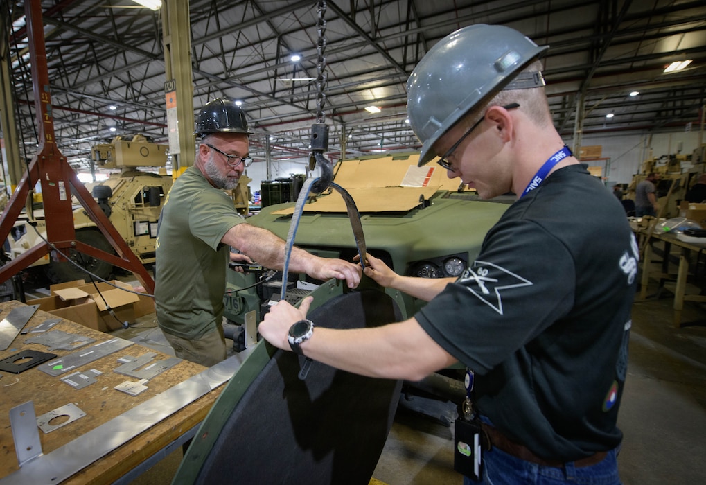 In a workshop, two workers use a hoist with straps to carefully attach a large panel to a green tactical vehicle.