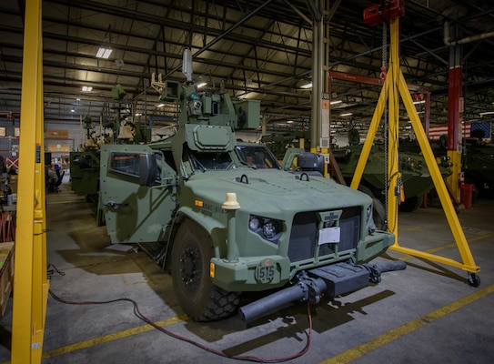 A green armored tactical vehicle sits inside a large factory, with a yellow hoist and other assembly equipment nearby.